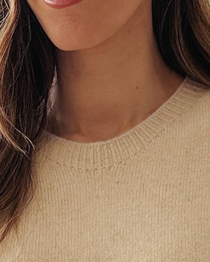 A close-up of a woman wearing a soft cream-colored knit sweater, with her hair and lower face visible against a bright indoor background.