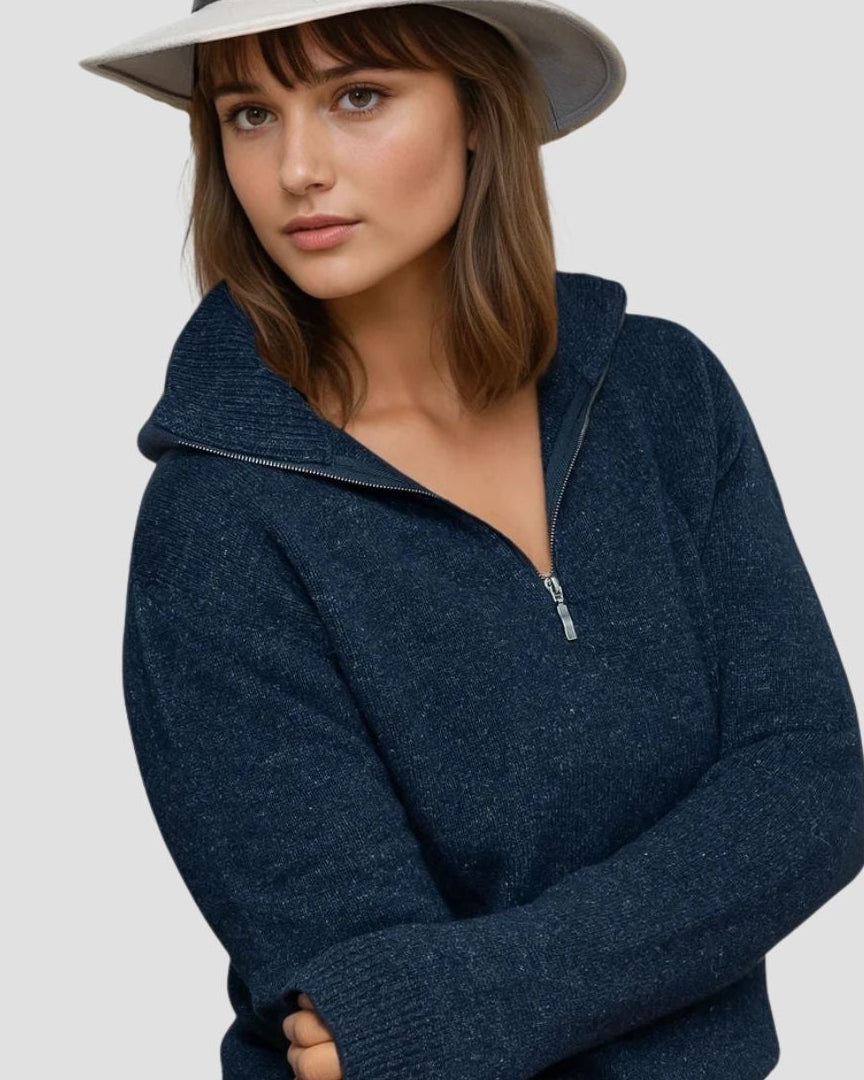 Woman wearing a navy blue zip-up sweater and white hat on a light gray background