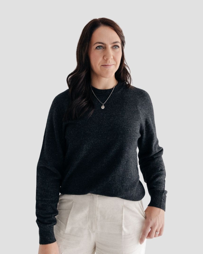 Woman wearing a dark gray sweater and light-colored pants against a plain background