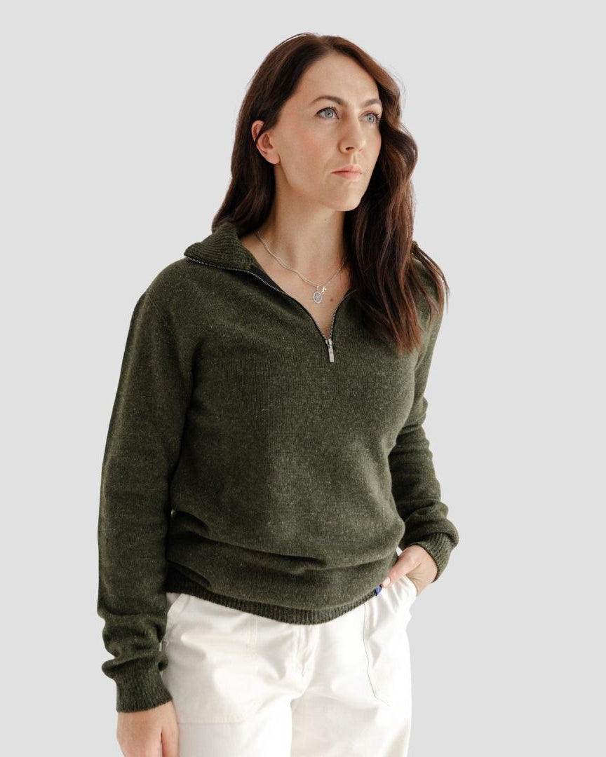Woman wearing a green sweater and white pants against a plain background