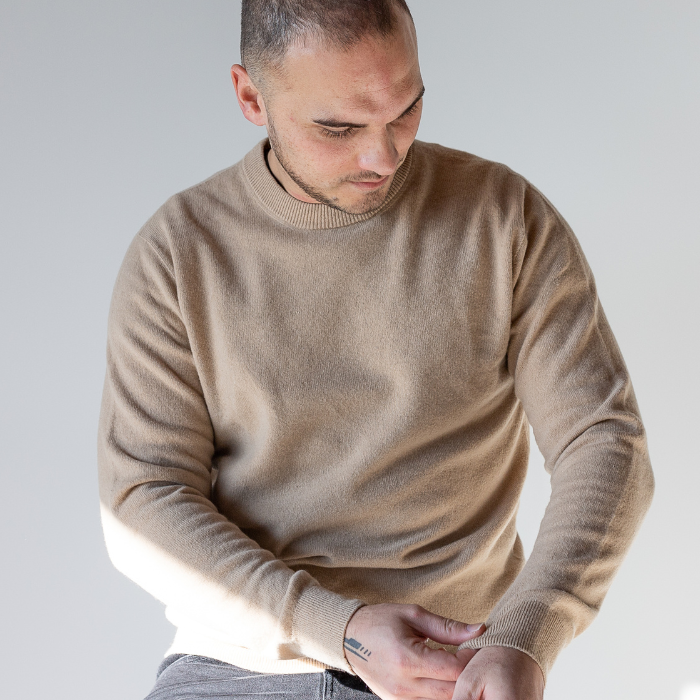 Man wearing unisex neutral toned crew neck jersey and looking at detailing on sleeve. 