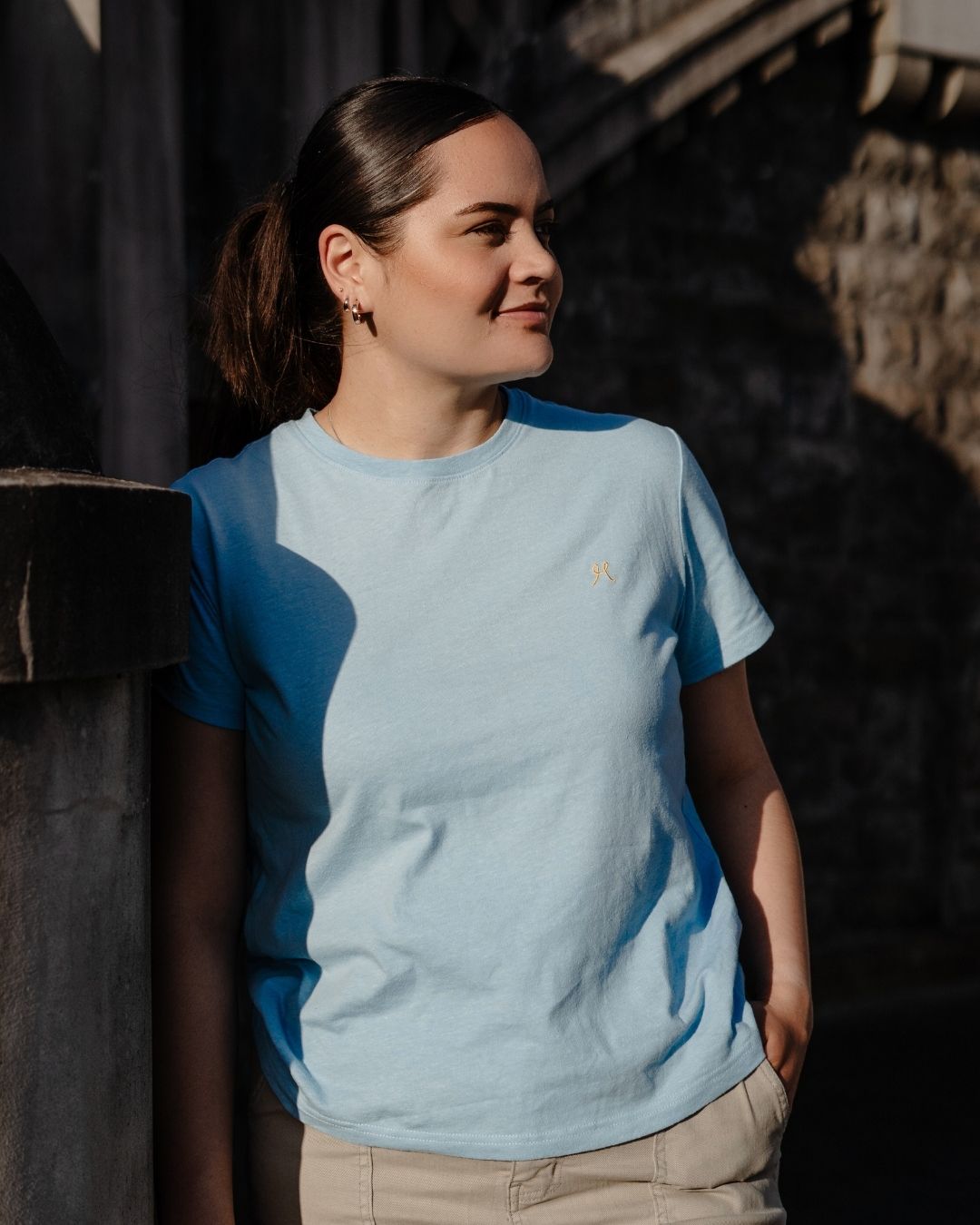 Woman wearing a light blue t-shirt with a small logo, standing against a dark background.