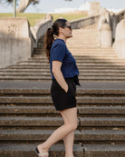 Woman in a blue shirt and black shorts standing on stone steps with a scenic background