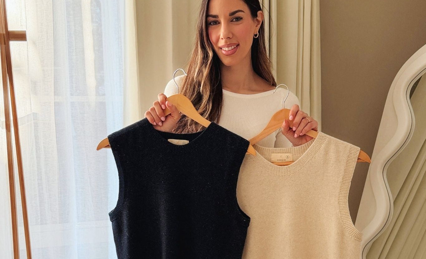 A woman holds up two sleeveless knit vests on wooden hangers, one black and one cream/beige, in a softly lit indoor setting with curtains and a mirror in the background.
