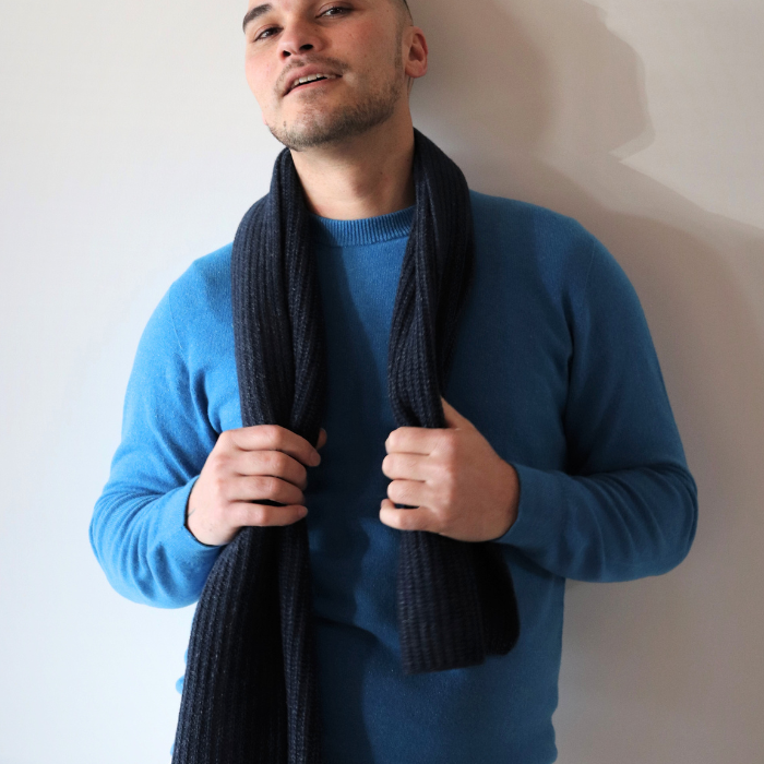 Man wearing a bright blue hemp and merino jersey and chunky knit scarf in charcoal. Leaning against wall in relaxed pose. 