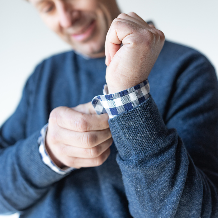 Middle aged man doing up cuff links on the sleeve of his shirt. Also wearing a navy crew neck sweater jersey which looks soft and tidy.