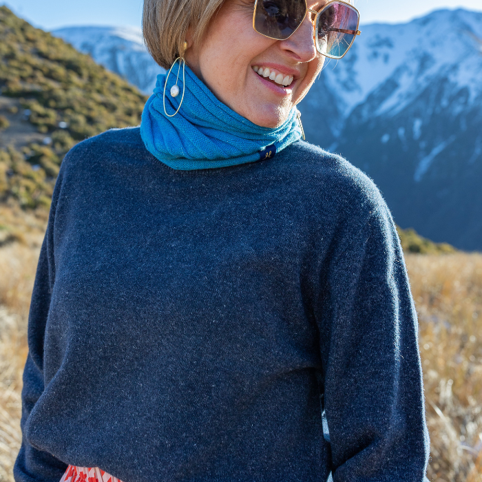 Middle aged women in navy crew neck sweater from Hemprino knitwear. Enjoy the mountains and snow behind her, possibly on a picnic. Also wearing a bright blue neck warmer or buff.
